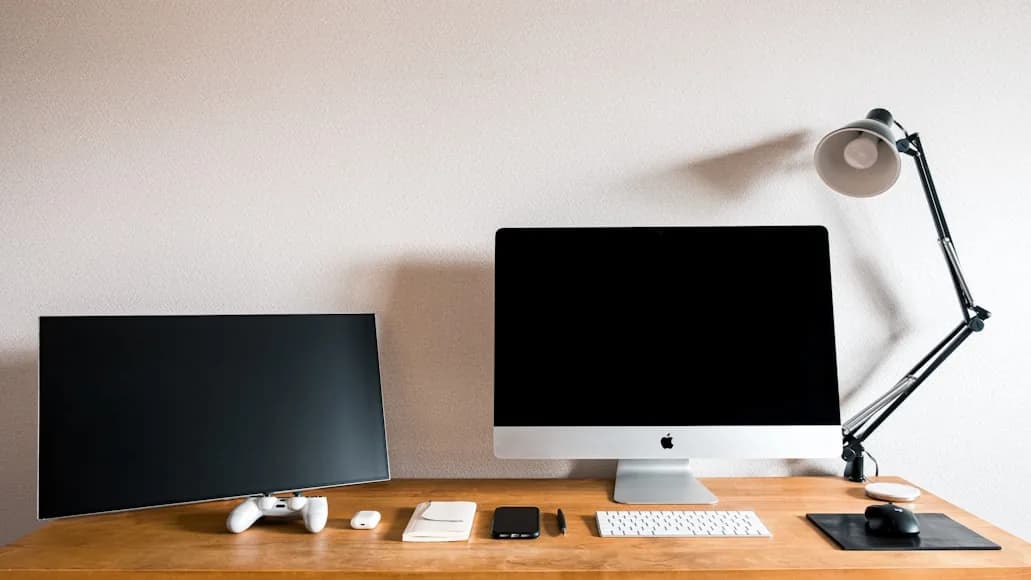 A compact wooden desk with an iMac, a secondary monitor, keyboard, mouse, and adjustable task lamp.