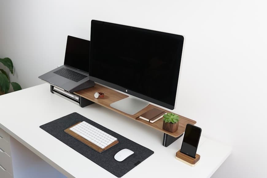 A white desk with a wooden monitor riser holding a desktop display and a laptop, with a keyboard, mouse, and phone arranged neatly below.