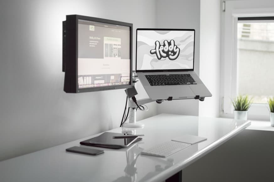 A white desk with an external monitor and an open laptop raised on adjustable arms beside a window.