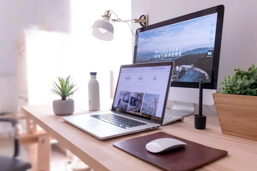 A wooden desk with an open laptop in front of a large monitor, a mouse pad, and a wall-mounted lamp in a compact two-screen setup.