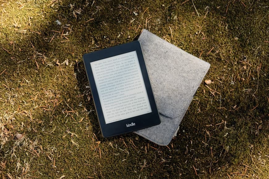 A Kindle e-reader resting on a desk beside a cup of coffee and a small plant, in a clean home workspace.