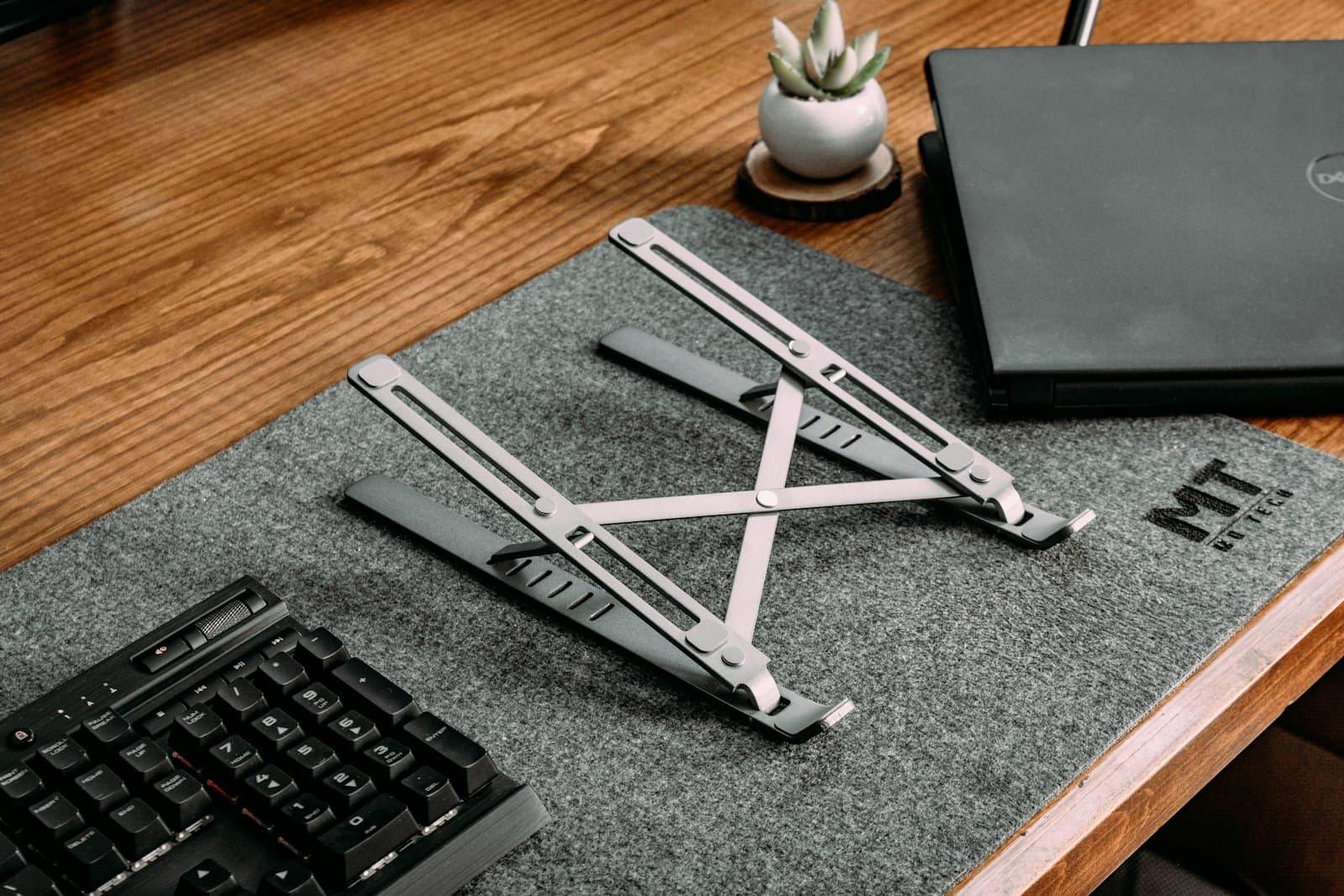 A foldable aluminum laptop stand on a gray desk mat beside a keyboard and a closed laptop on a wooden desk.