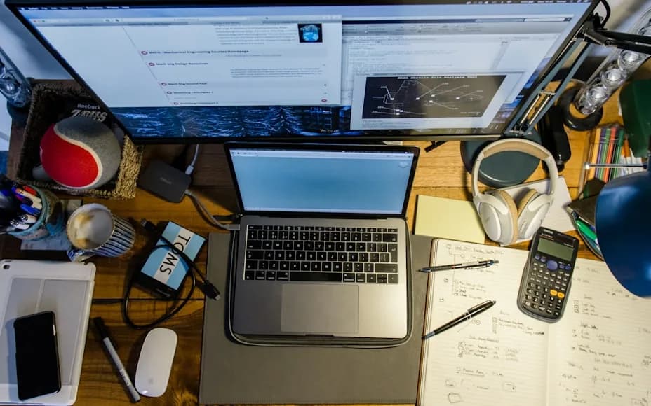 An overhead view of a desk with dual monitors, a laptop, notes, headphones, and desk accessories.