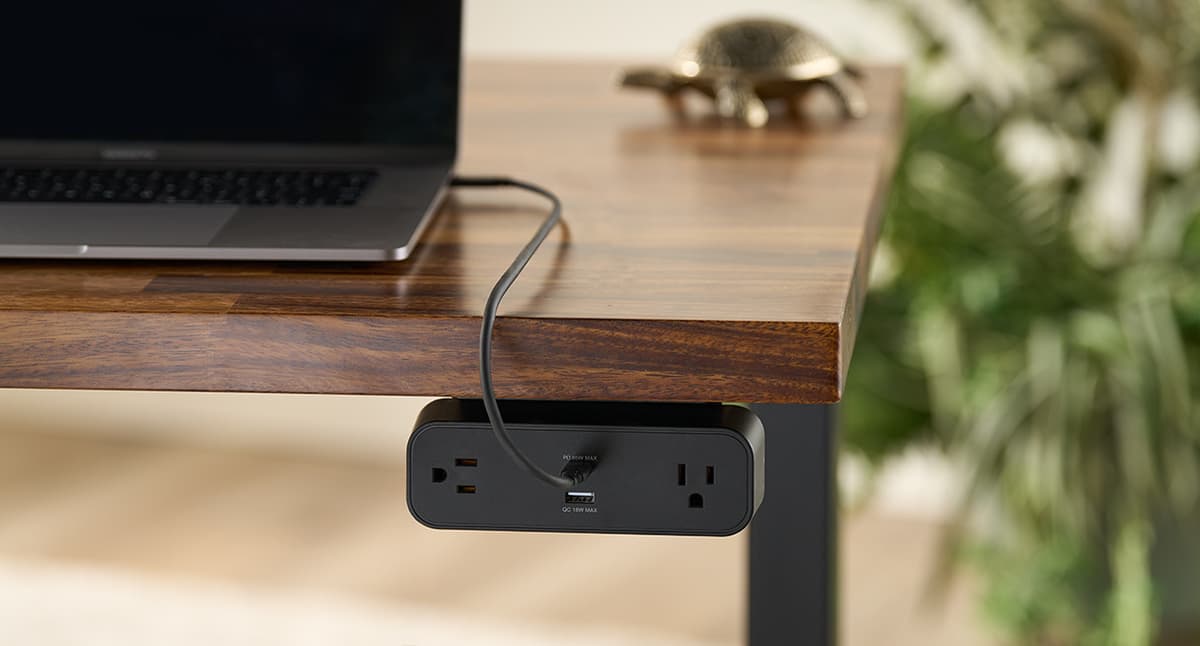 A black power strip mounted beneath the front edge of a wood desk with a charging cable running across the desktop.