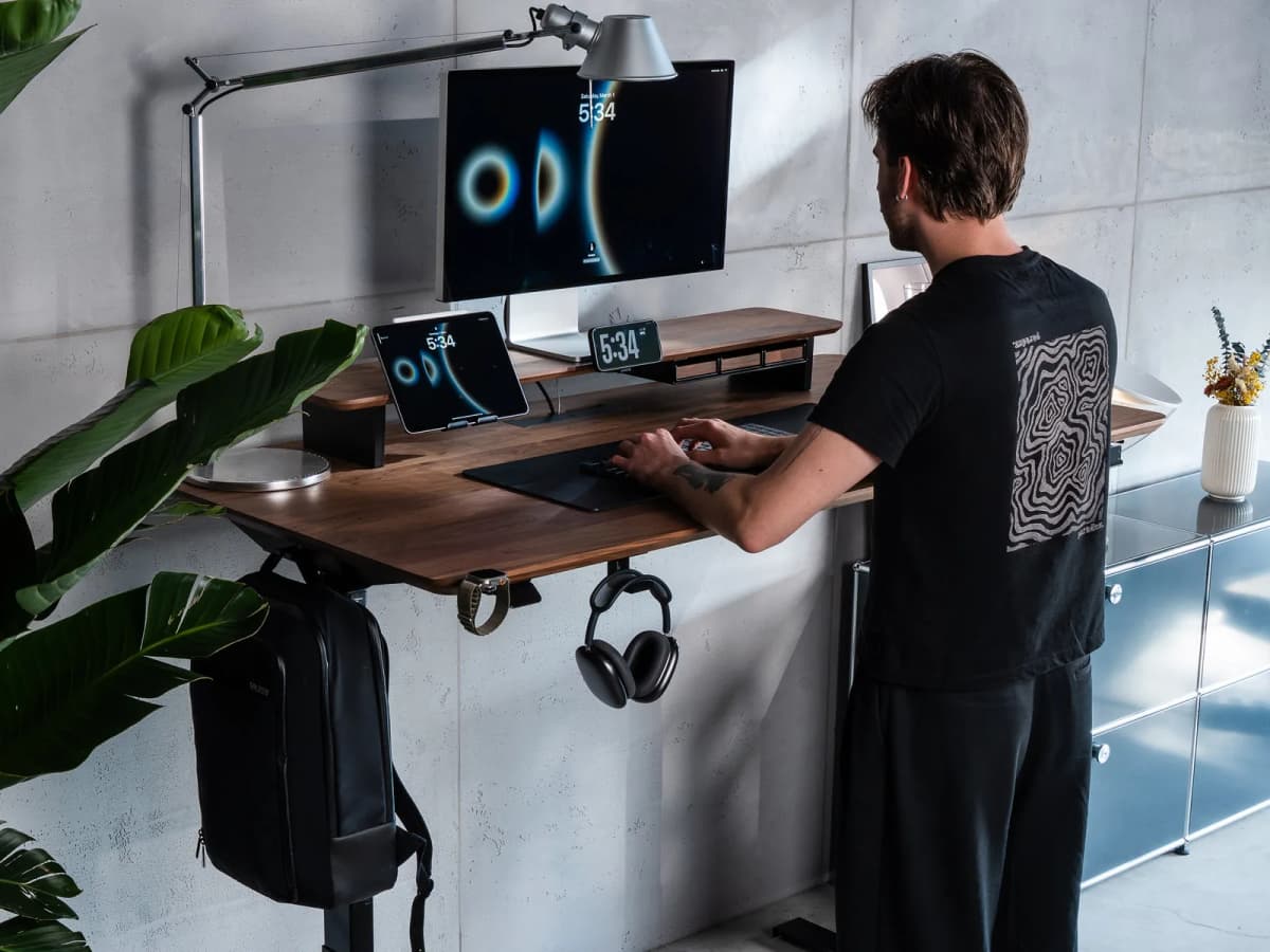 A pair of over-ear headphones hanging from a slim holder mounted under a desk shelf beneath a monitor.