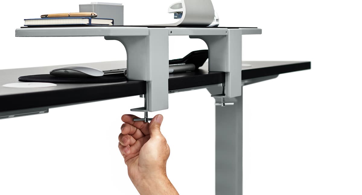 The 15-inch UPLIFT desk shelf clamped to a desk edge with accessories stored on top.