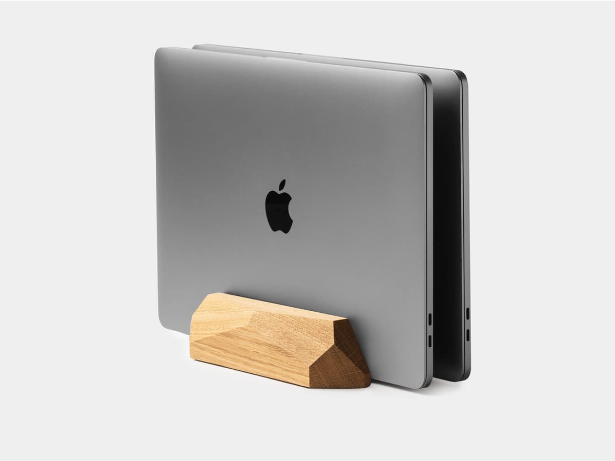 A closed laptop resting in an Oakywood wooden vertical dock on a desk beside a monitor.
