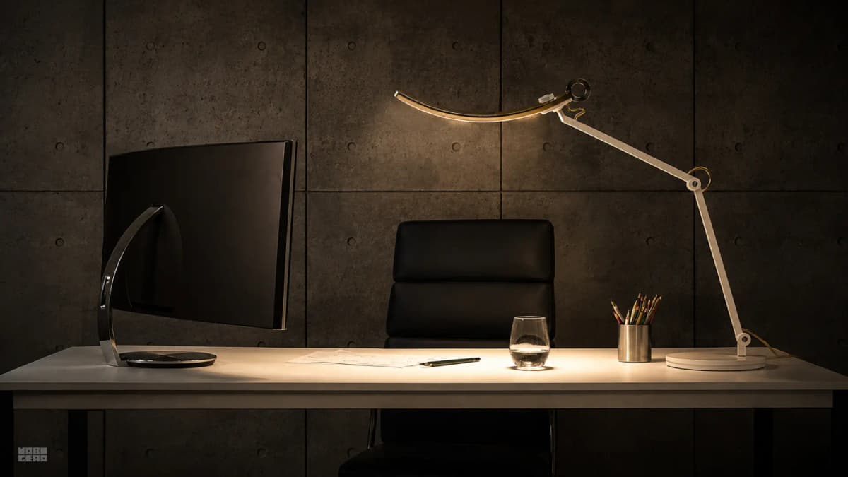 A modern desk with a curved monitor, an articulated desk lamp casting warm light, and a black leather chair against a dark concrete wall.