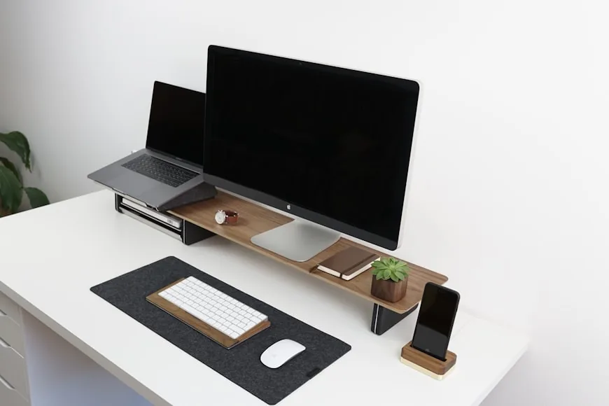 A white desk with a wooden monitor riser holding a desktop display and a laptop, with a keyboard, mouse, and phone arranged neatly below.