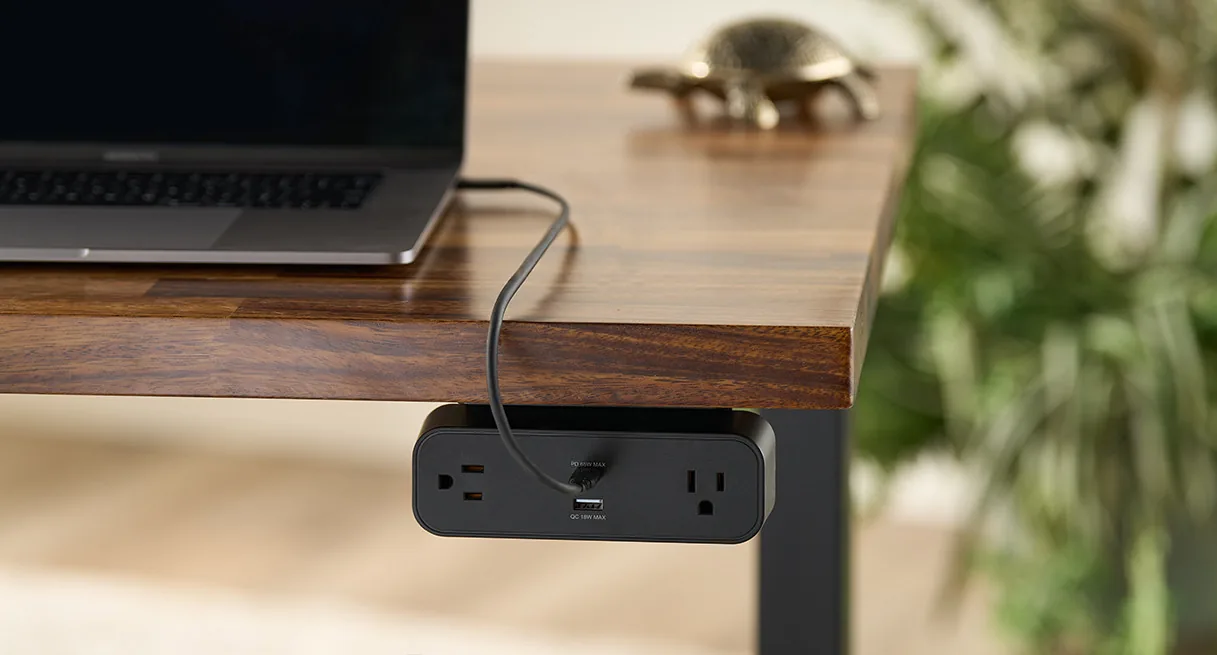 A black power strip mounted beneath the front edge of a wood desk with a charging cable running across the desktop.