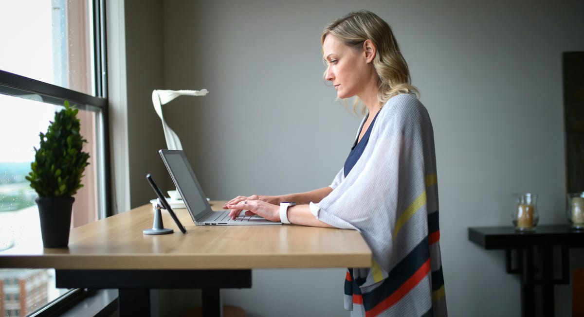 Woman working at a height-adjustable desk beside a window with a laptop.