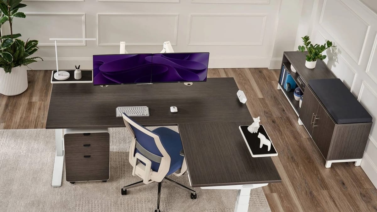 High-angle overview of an L-shaped standing desk workstation with dark ash top, white 3-leg frame, and dual displays in an executive home office.